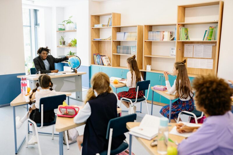 a man in black suit talking to his students while holding a globe on the table